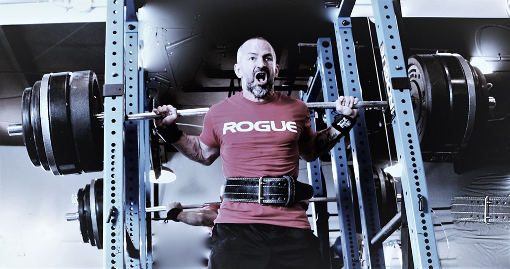 S2 stew squatting-min S2 Method Stewart Breeding in red Rogue brand tshirt performing squats with heavy weight on the bar