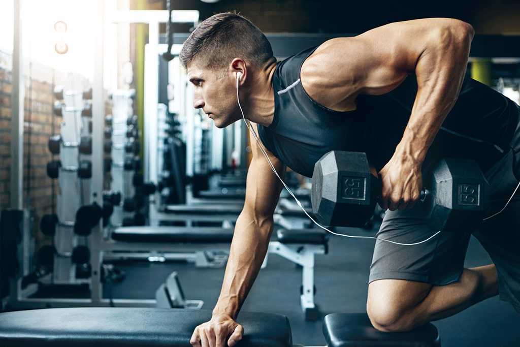 S2 Method Meditraining Medilifting Man at gym performing one arm row while kneeling on bench and listening to audio through earbuds