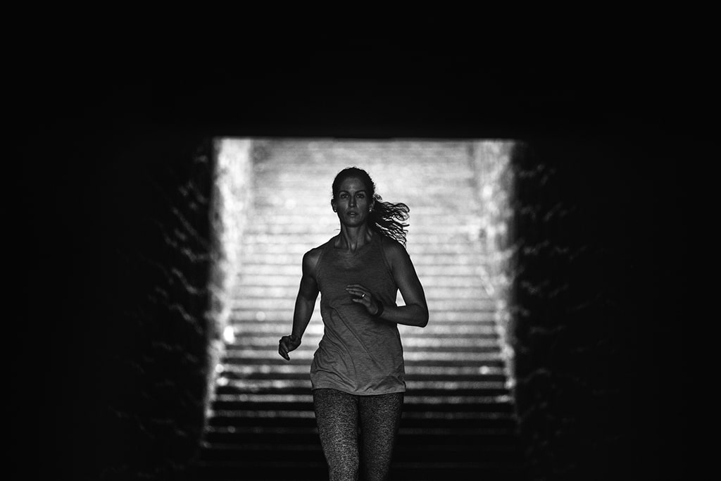 An adult woman training on a large set of outdoor steps, running and employing other stairway exercise variations. High contrast black and white image of her running through a tunnel at the bottom of the stairway.